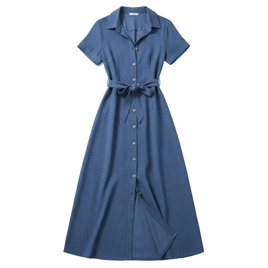 A woman models a blue short-sleeve maxi dress with a button-down front, a tie waist, and a high leg slit. She is standing outdoors against a rustic wooden door and a white stone wall, holding a brown woven handbag.