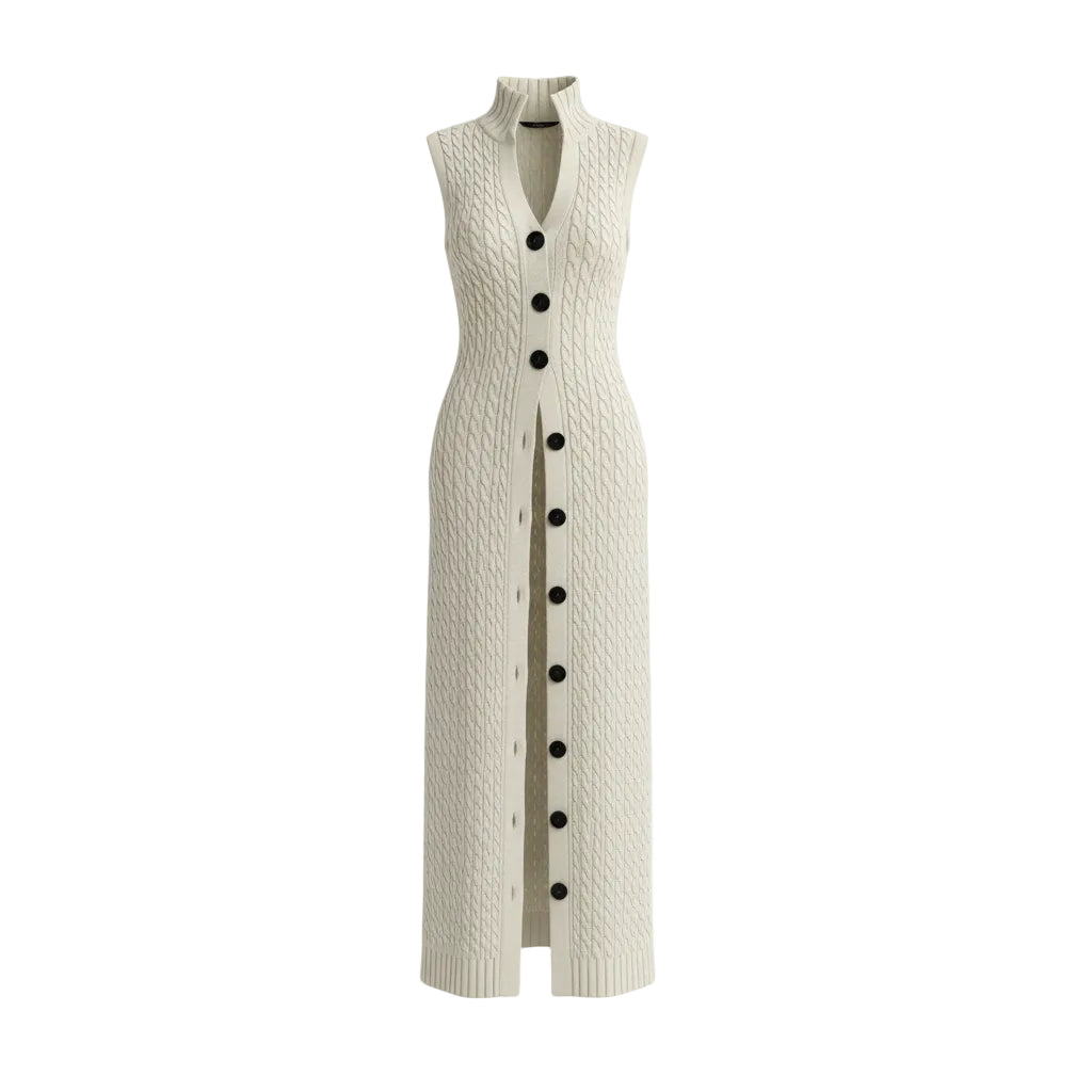A woman models a long cream cable-knit sleeveless cardigan vest with large black buttons, paired with white wide-leg pants and holding a woven beige handbag against a textured gray studio background.